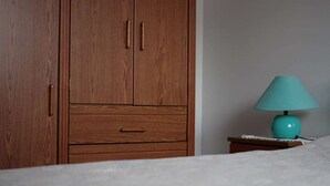 Une chambre avec une armoire en bois, un seul tiroir, une lampe turquoise sur une table de chevet et un lit recouvert d’une couette blanche.