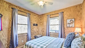 A bedroom with a wooden wall, a bed with a checkered bedspread, two windows with curtains, a ceiling fan, and a lamp on the nightstand. - 5 Mi to Downtown Morganton: Cabin on Horse Farm (Morganton)