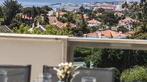 A dining table set with plates, glasses, and a decorative vase, overlooking a coastal town with buildings and the sea. - Estoril Queen's Apartment by CadenzaLux (Estoril)