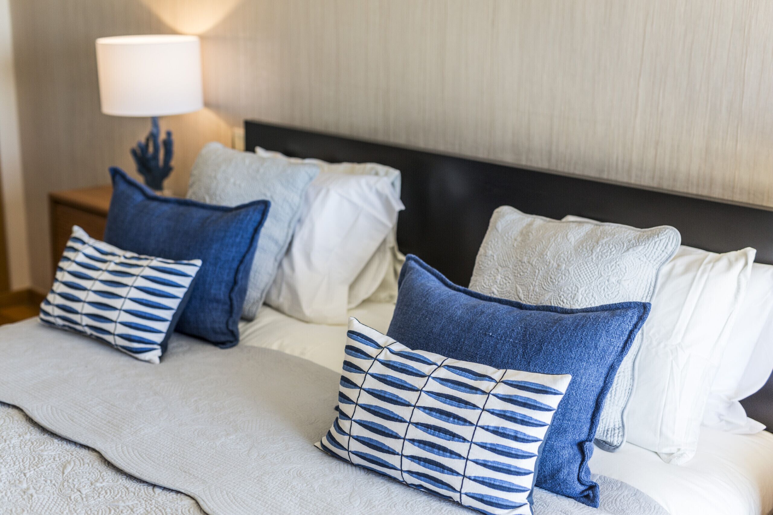 Ein ordentlich bezogenes Bett mit einer weißen und blauen Decke sowie Kissen, ein Nachttisch mit Lampe und ein Kopfteil mit strukturierter Gestaltung.