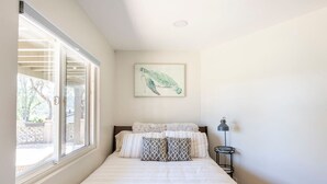 Ein Schlafzimmer mit großem Fenster, einem Bett mit gemusterten Kissen, einem Wandbild und einem Nachttisch mit Lampe.