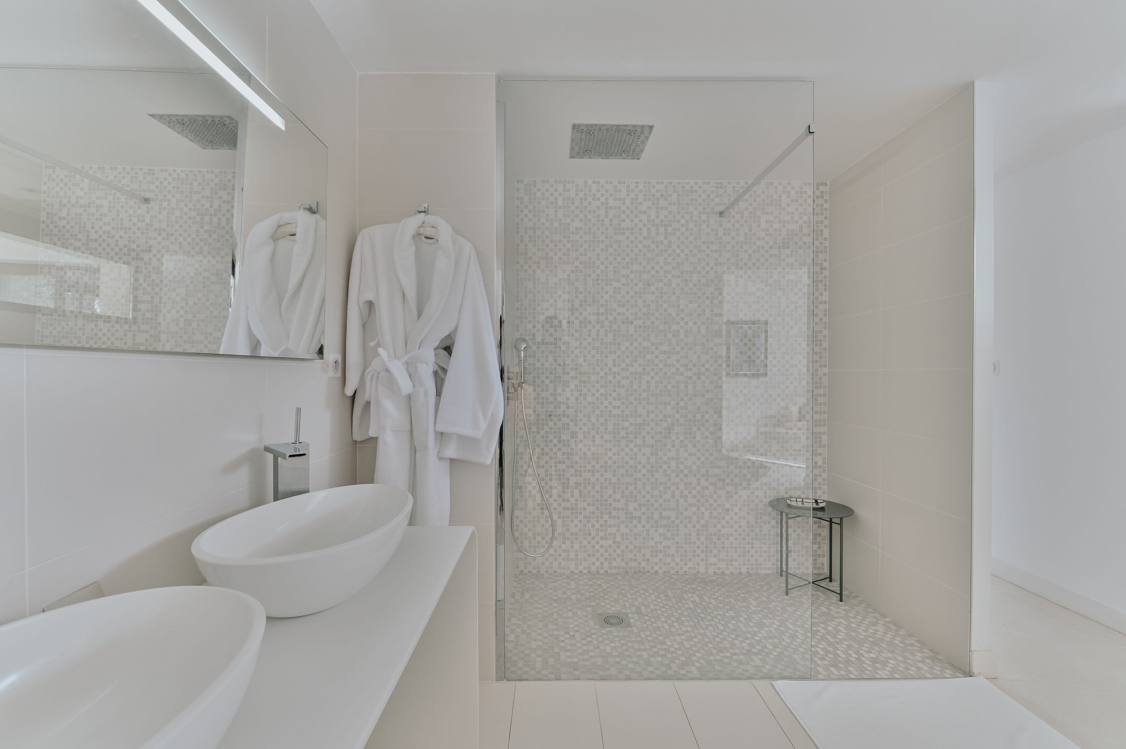 Een moderne badkamer met een douchecabine van glas, een groot wit wastafel en twee witte badjassen hangend aan haken.
