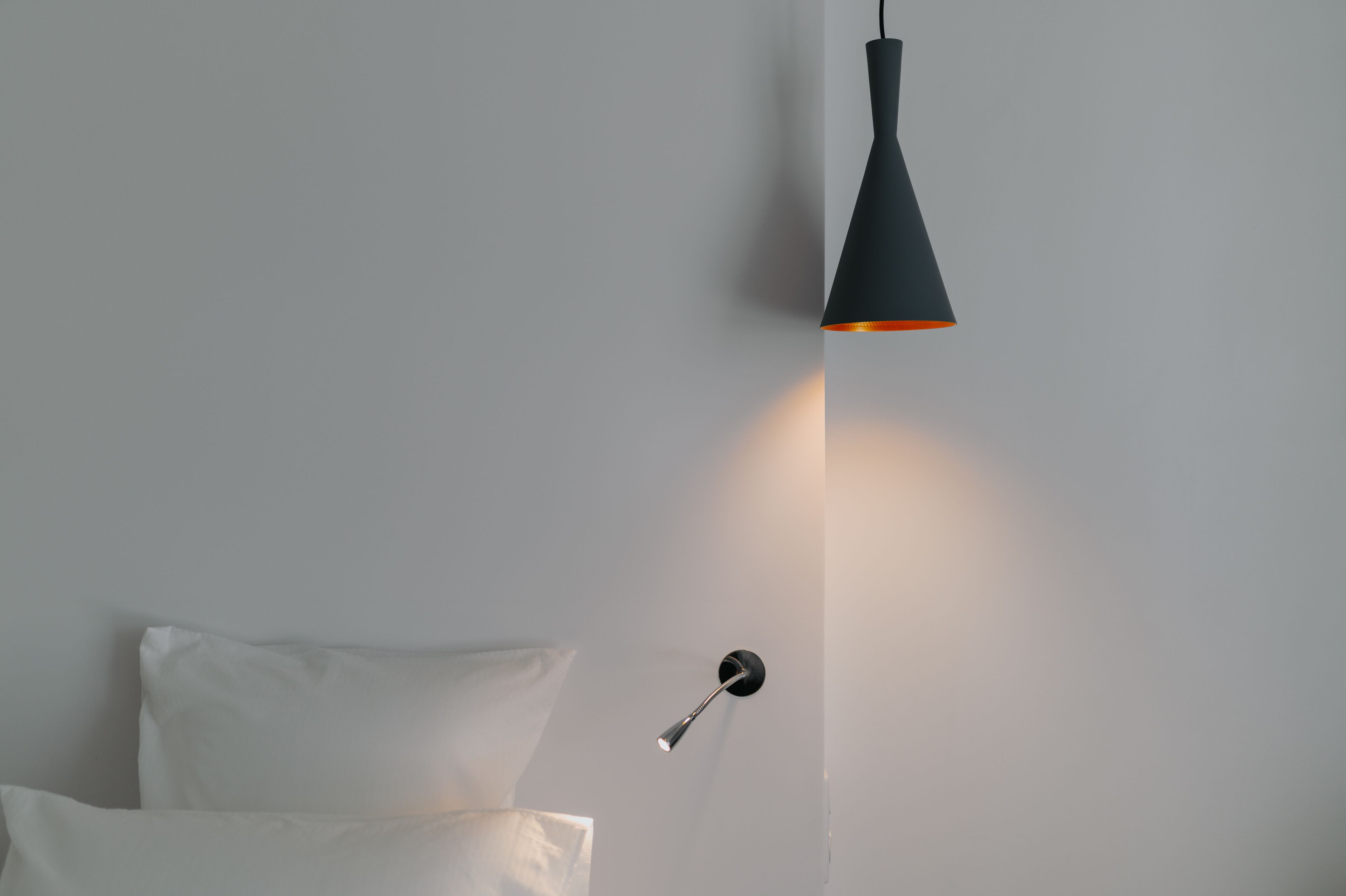 A modern room with a minimalist design, featuring a single bed with white bedding and a wall-mounted light fixture.