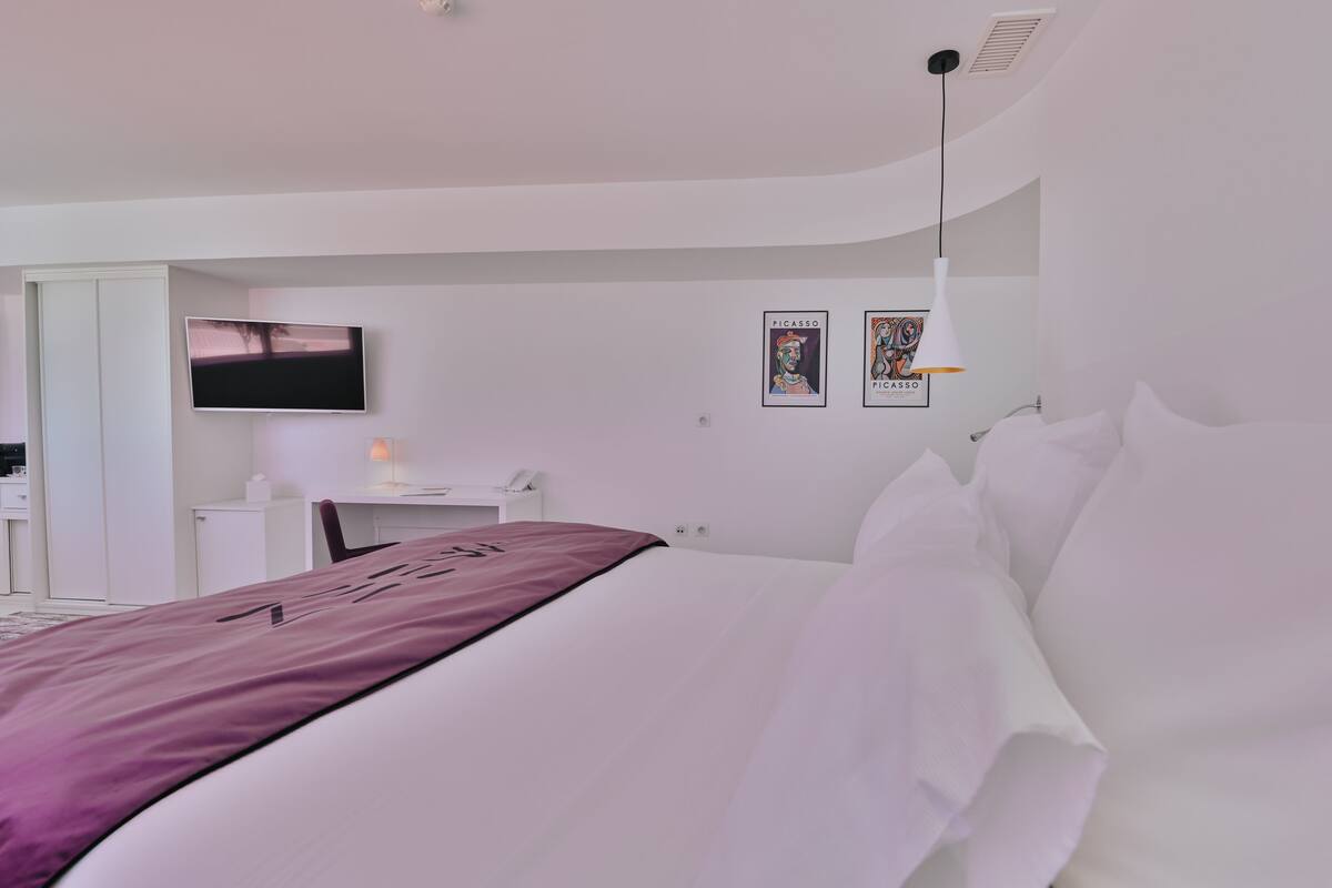 A modern hotel room with a bed, a desk, a TV, and artwork on the wall.