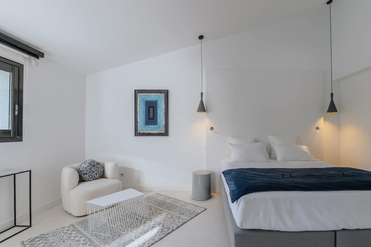 A modern bedroom with a bed, bedside lamps, a chair, a small table, and a decorative wall piece.