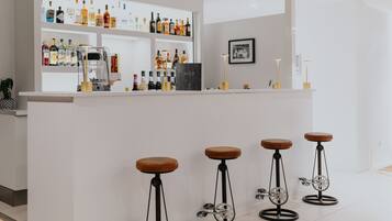 Eine Barbereich mit einer weißen Theke, Regalen voller Spirituosenflaschen, einer Kaffeemaschine und vier Ketten-Hockern.