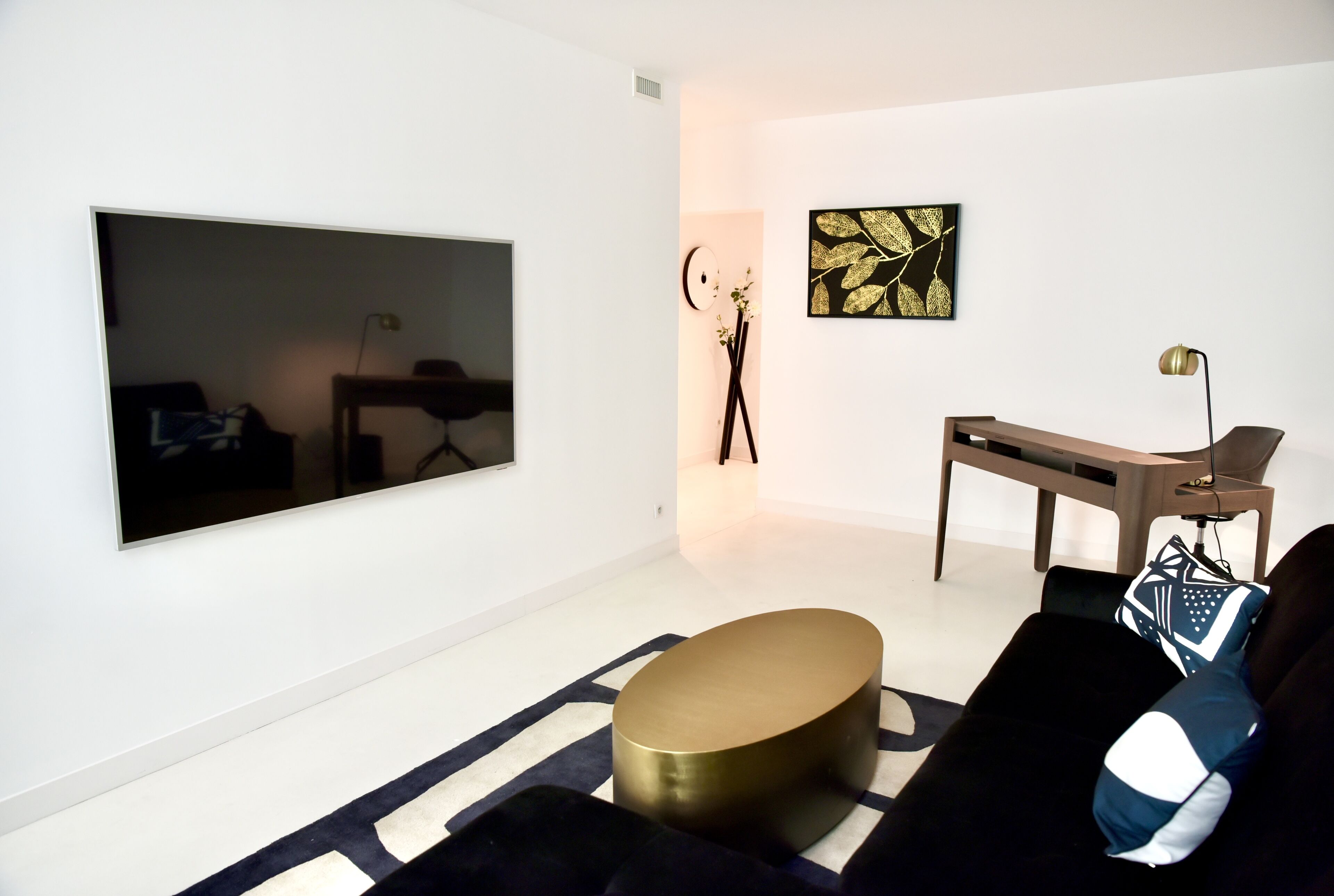 Una sala moderna con un televisor de pantalla plana grande montado en la pared, una mesa de centro redonda dorada, un sofá negro con cojines estampados y una mesita auxiliar minimalista.