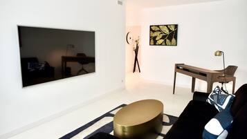 Una sala moderna con un televisor de pantalla plana grande montado en la pared, una mesa de centro redonda dorada, un sofá negro con cojines estampados y una mesita auxiliar minimalista.