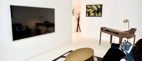 Una sala moderna con un televisor de pantalla plana grande montado en la pared, una mesa de centro redonda dorada, un sofá negro con cojines estampados y una mesita auxiliar minimalista.