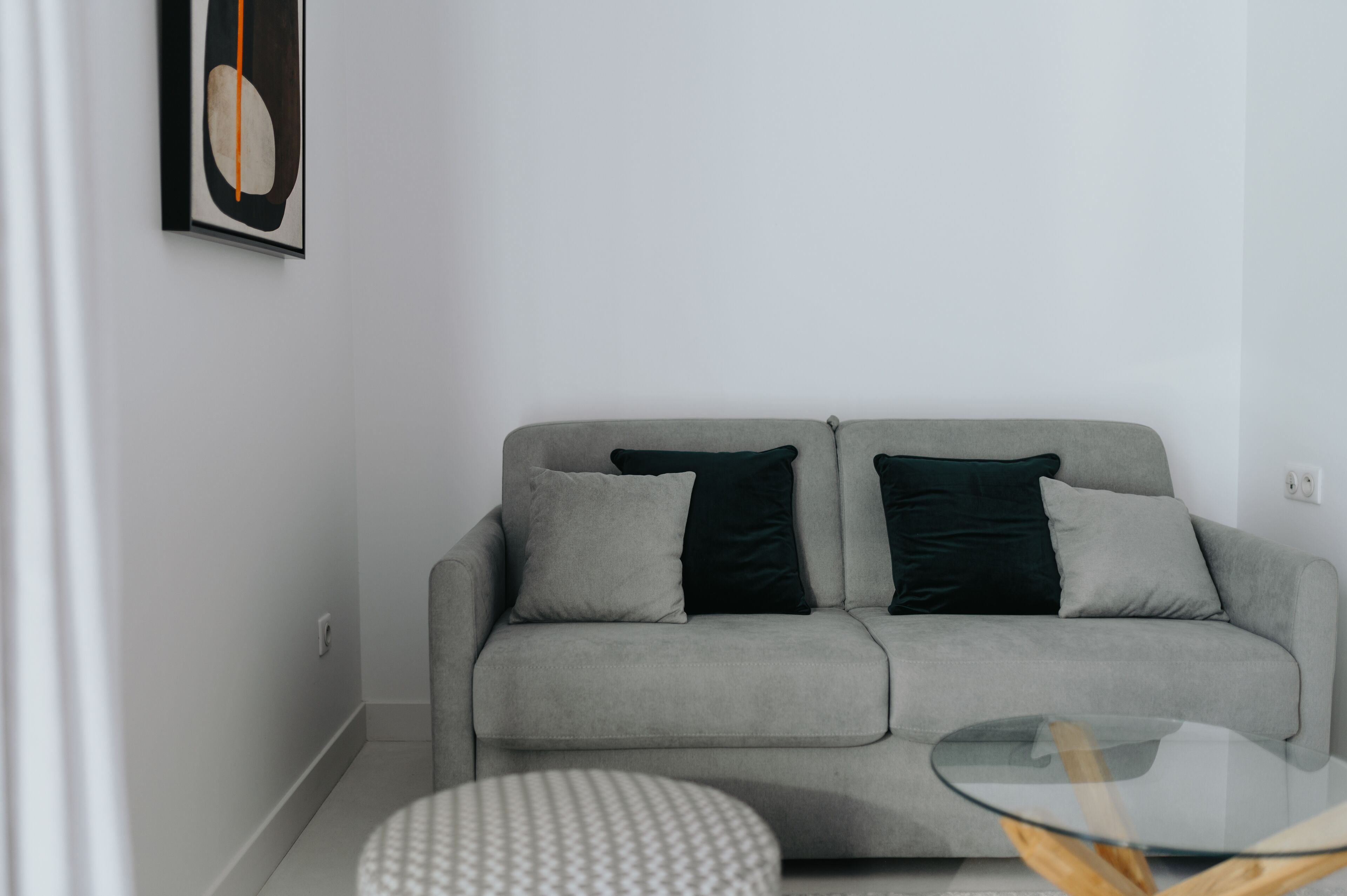 A modern living room with a grey sofa, two black cushions, a glass coffee table, and a patterned ottoman.