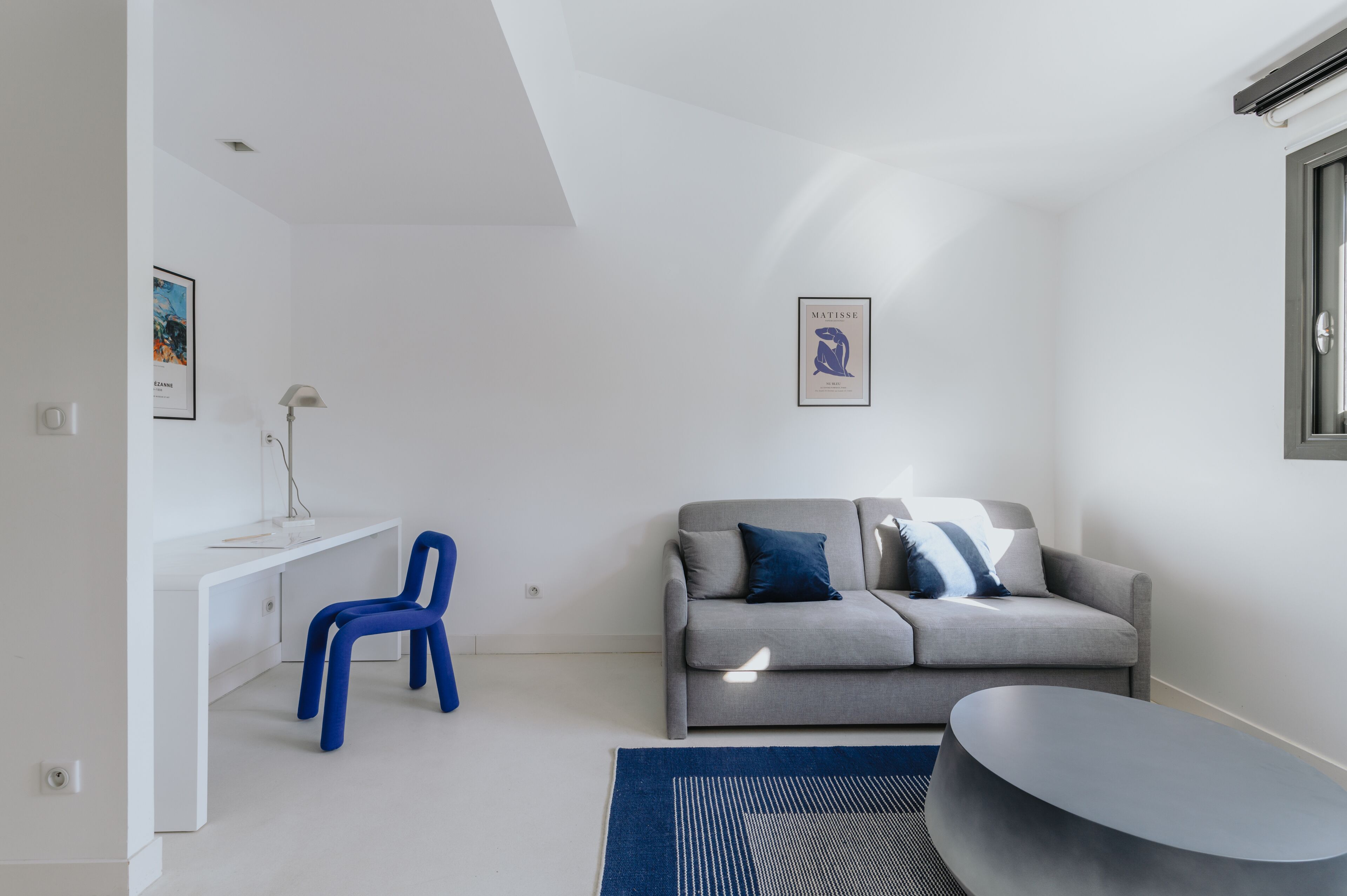 Una sala de estar moderna y minimalista con un sofá gris, un sillón azul y un escritorio blanco.