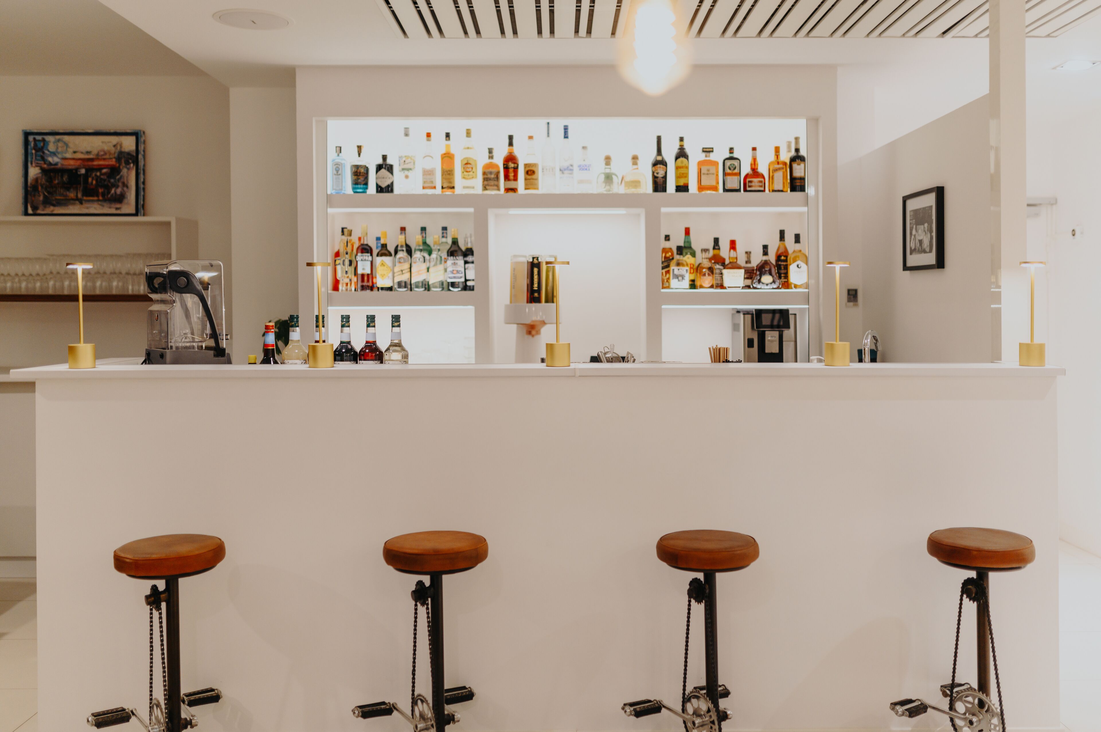 Zona de bar con mostrador blanco, taburetes de madera y estantes llenos de botellas diversas.