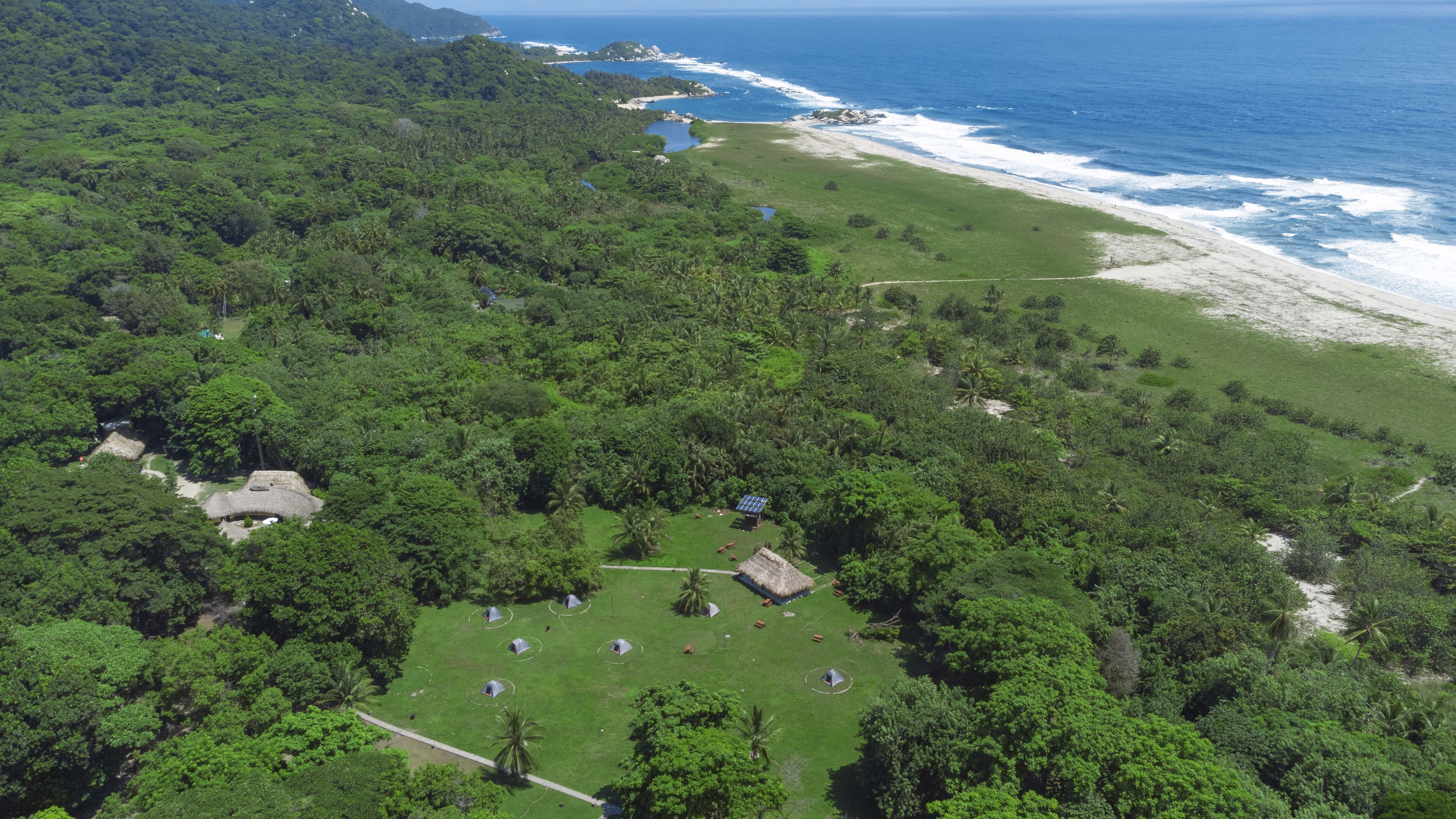 Vue aérienne d’un complexe hôtelier tropical doté de plusieurs bungalows aux toits de chaume, d’une plage et d’une forêt dense.