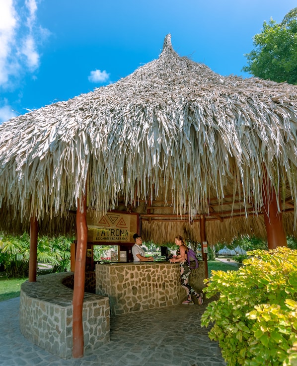A thatched-roof structure with a stone base, a sign reading "AURONA COHAB&SABANAS", and a person standing at the counter.