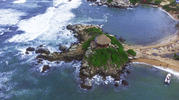 Vista aérea de una zona costera con un promontorio rocoso, una cabaña con techo de paja y una playa de arena con gente.