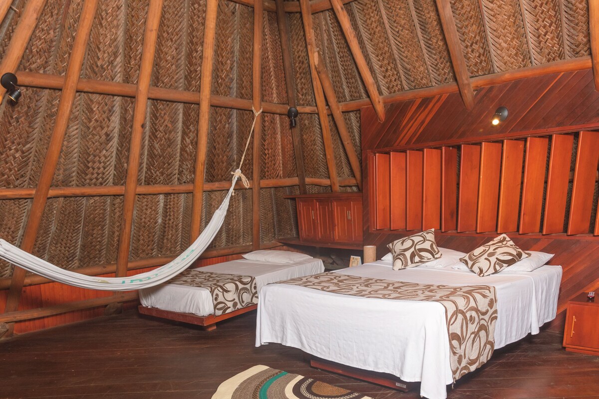 Una habitación con dos camas, una hamaca, paredes de madera y techo tejido.