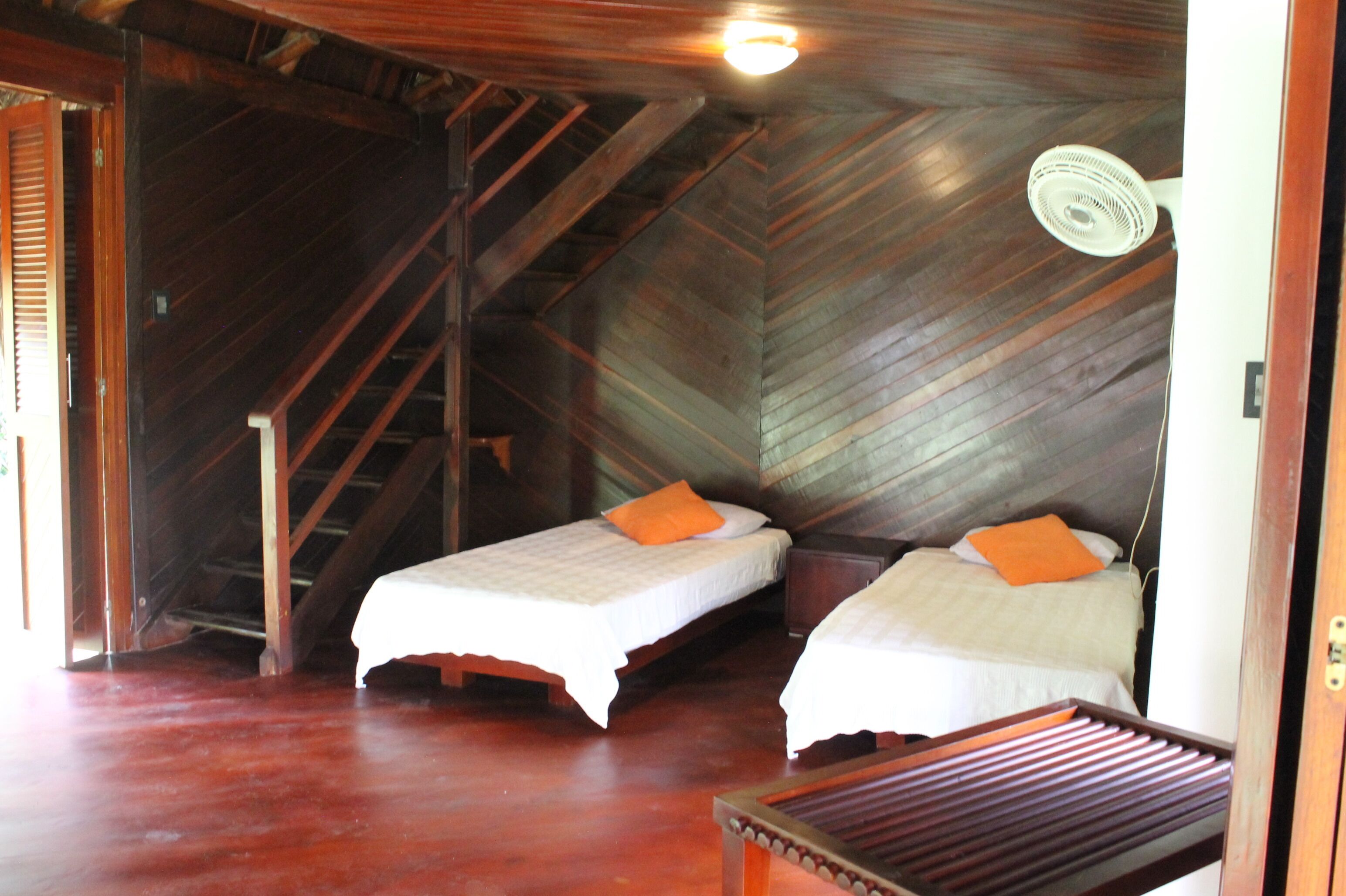 A room with two beds, wooden flooring, a staircase, and a ceiling fan.