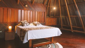 Un dormitorio con techo inclinado de madera, una cama con ropa de cama estampada, una mesita de noche con una lámpara y una alfombra tejida.