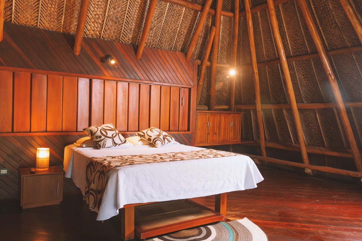 A bedroom with a wooden sloped ceiling, a bed with patterned bedding, a nightstand with a lamp, and a woven rug.