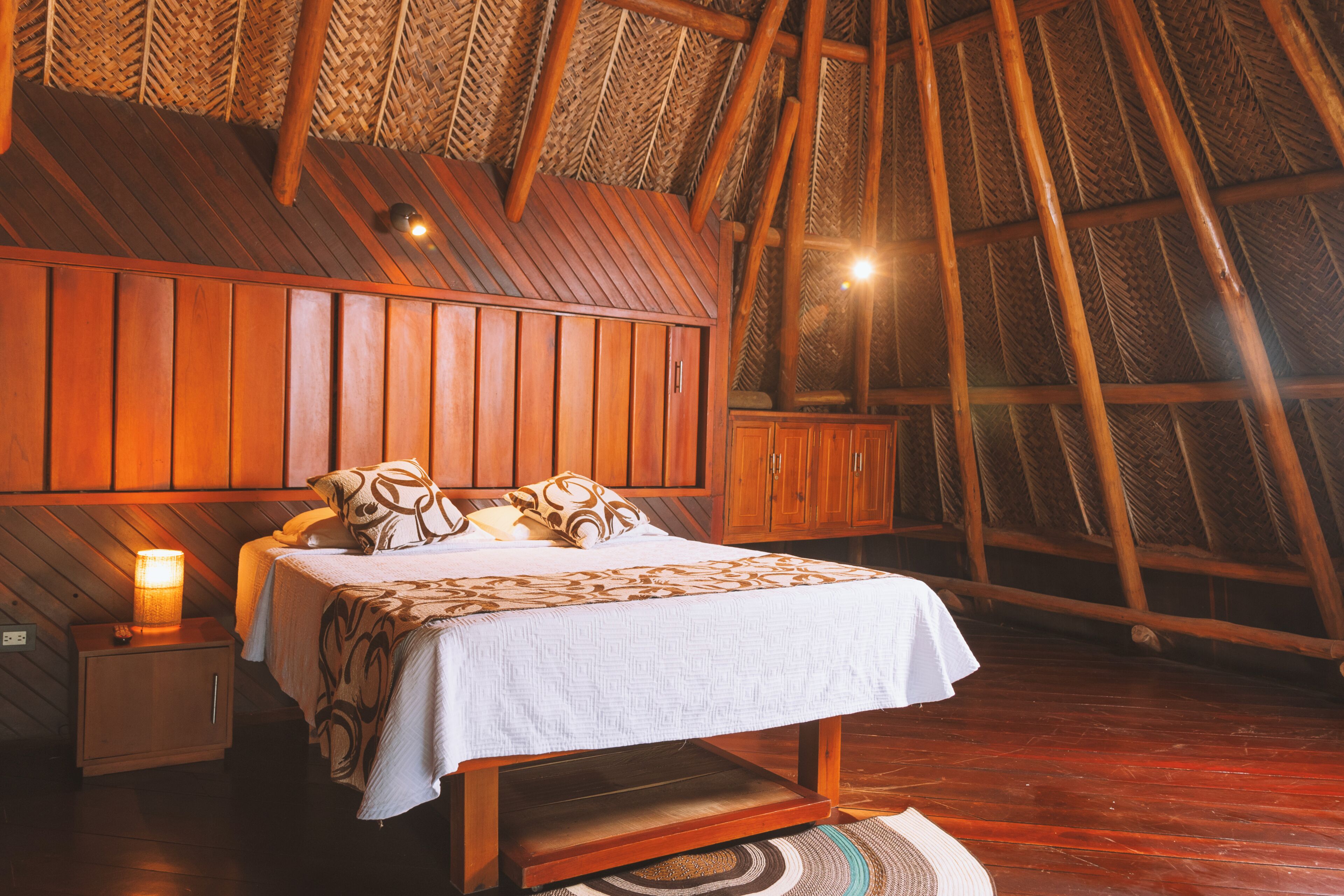 Une chambre avec un plafond en pente en bois, un lit avec une literie à motifs, une table de chevet avec une lampe et un tapis tissé.