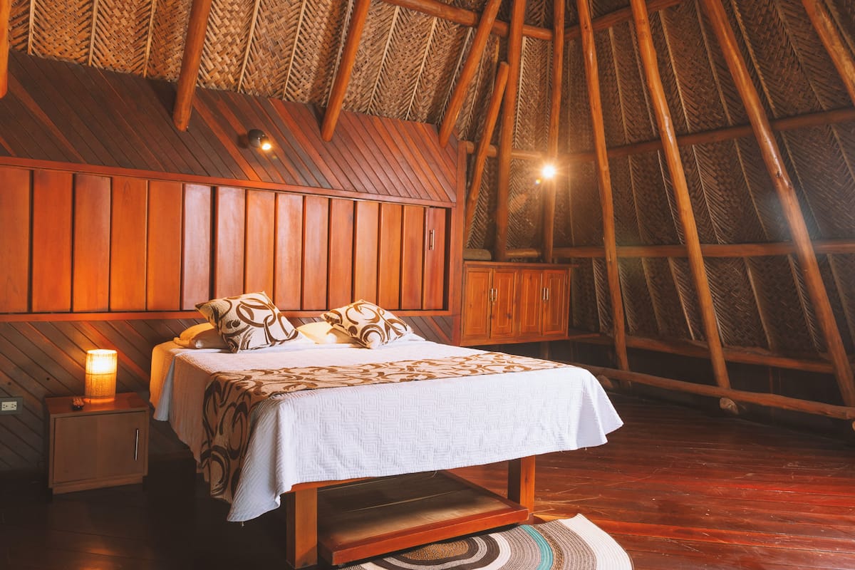A bedroom with a wooden sloped ceiling, a bed with patterned bedding, a nightstand with a lamp, and a woven rug.