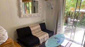 A room with a black sofa, a wooden side table, a patterned rug, a ceiling with wooden beams, a mirror with a decorative frame, and a sliding glass door leading to an outdoor area. - Seahorse:1 Queen Bed (East End)