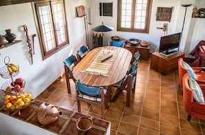 A dining area with a wooden table, chairs, and a rug. There are windows, a television, and various decorative items on the walls.