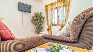 Ein gemütliches Wohnzimmer mit einer Couch, einem kleinen Tisch mit dekorativer Platte, einer Topfpflanze und einem an der Wand montierten Fernseher.