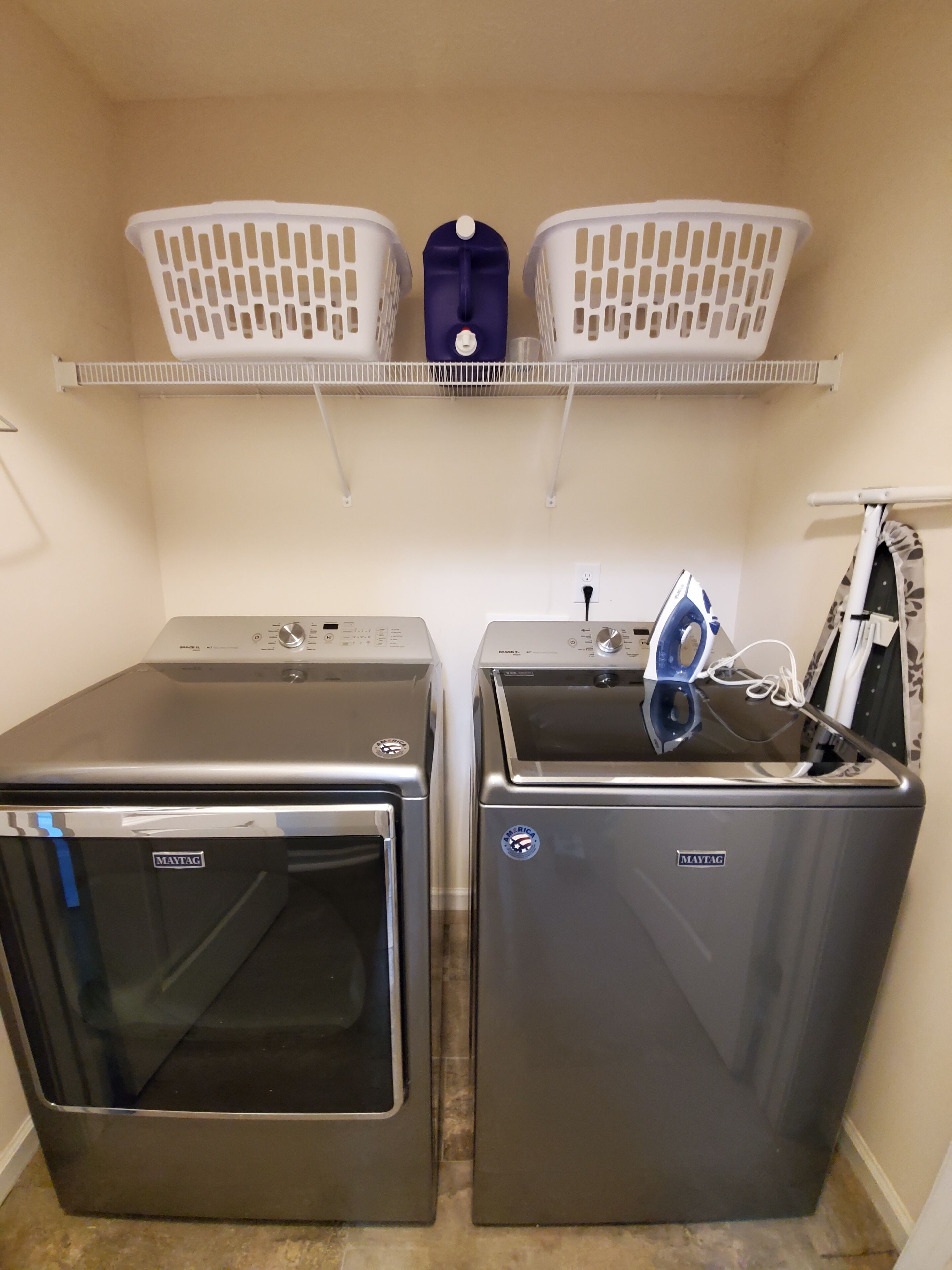 A laundry room with two Maytag washers and dryers, a blue iron, and two white laundry baskets on a shelf.
