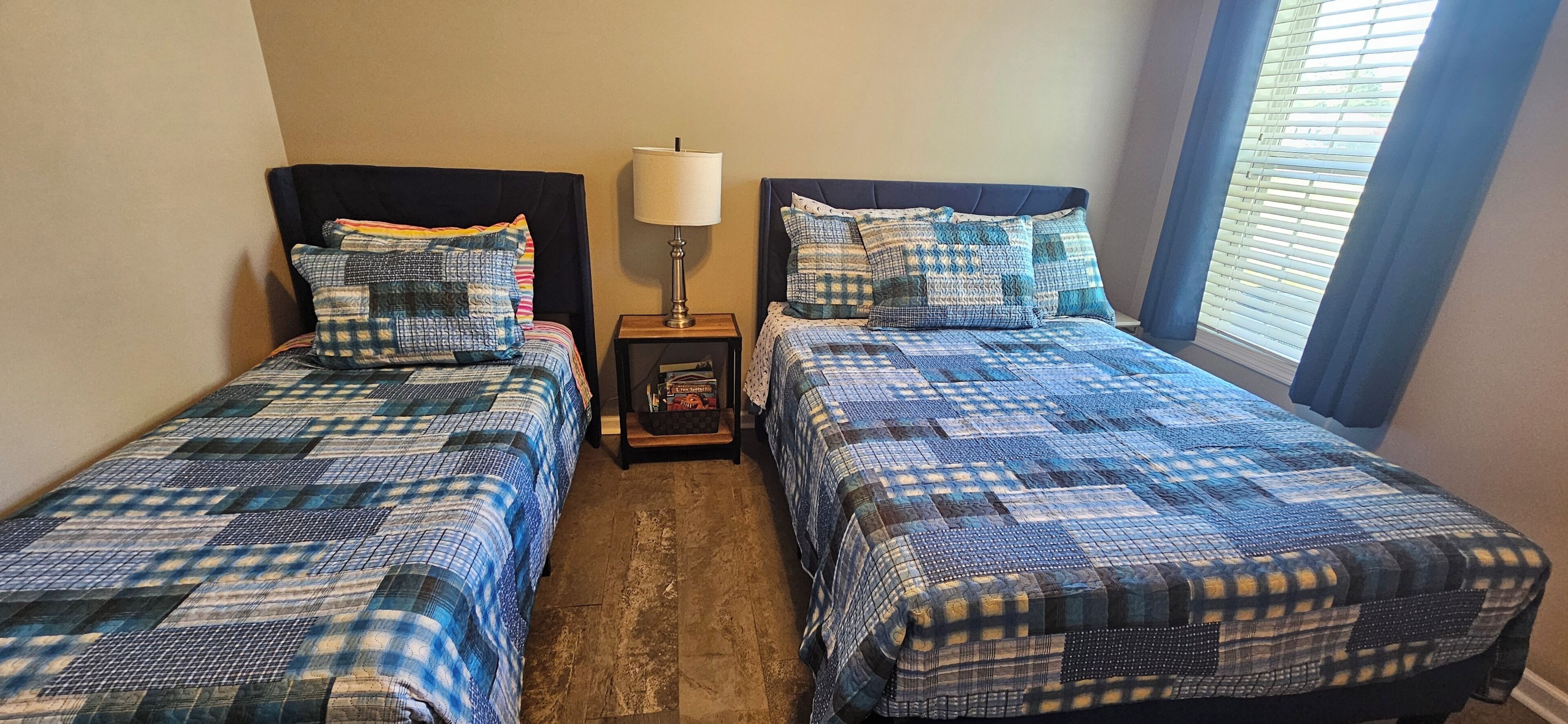 Dos camas individuales con ropa de cama a cuadros azul y negra a juego, una mesita de noche con lámpara y una ventana con persianas.