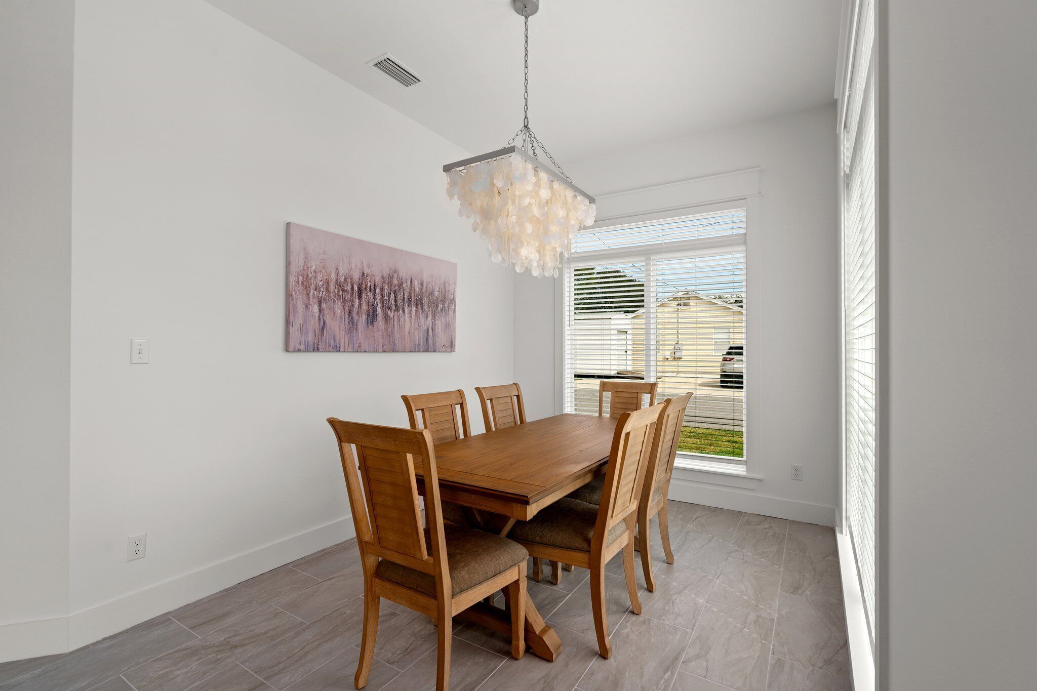 Een eetruimte met een houten tafel en stoelen, een hanglamp, een schilderij aan de muur en een raam met zonwering.