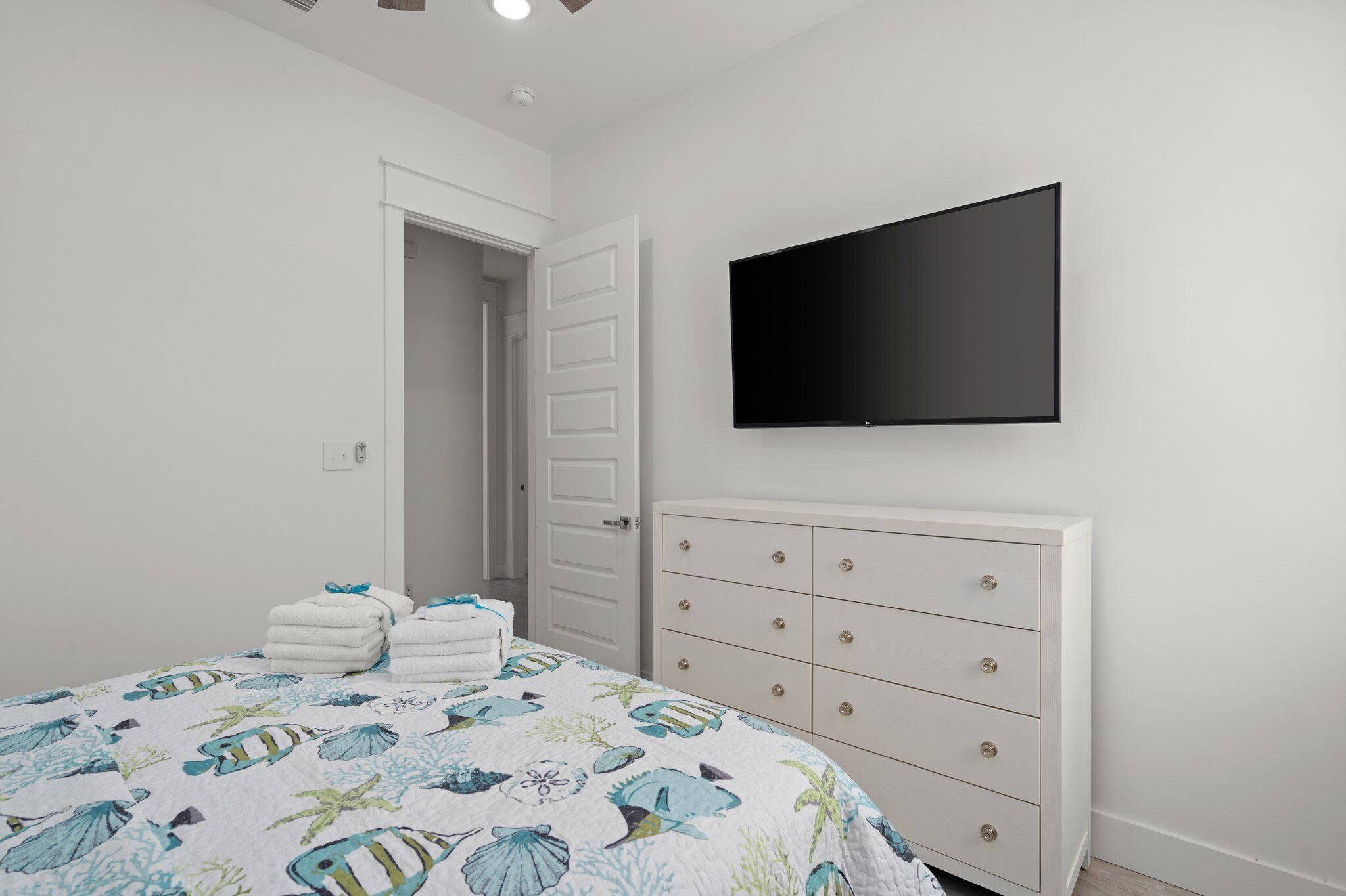 A bedroom with a bed, a white dresser, a flat-screen TV, and a door leading to another room.