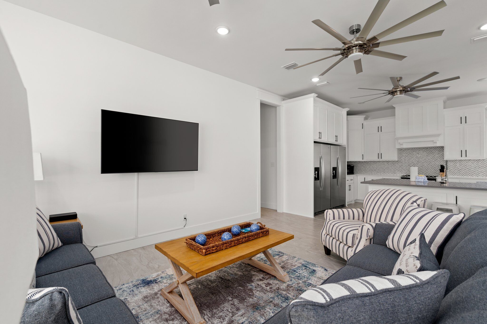 A modern living room with a flat-screen TV, a wooden coffee table with decorative items, a gray sofa, striped armchairs, and a ceiling fan.