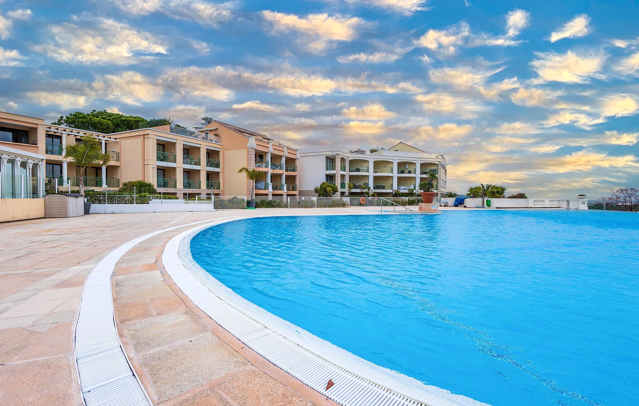 A large swimming pool with a curved edge, surrounded by a paved walkway. In the background, there are multi-story buildings with balconies and a clear sky with clouds.