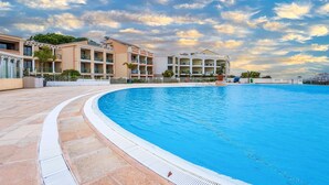 A large swimming pool with a curved edge, surrounded by a paved walkway. In the background, there are multi-story buildings with balconies and a clear sky with clouds. - Awesome apartment in Cannes with swimming pool (Cannes)