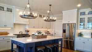 A modern kitchen with white cabinets, a stainless steel refrigerator, a large island with stools, and pendant lighting.