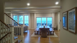 A spacious living area with a wooden staircase, a dining table with chairs, and large windows offering a view of the ocean.