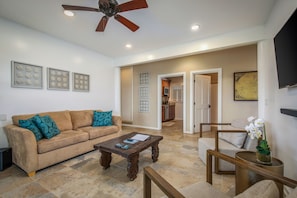 A living room with a sofa, coffee table, and a TV on the wall. There is a ceiling fan and recessed lighting.