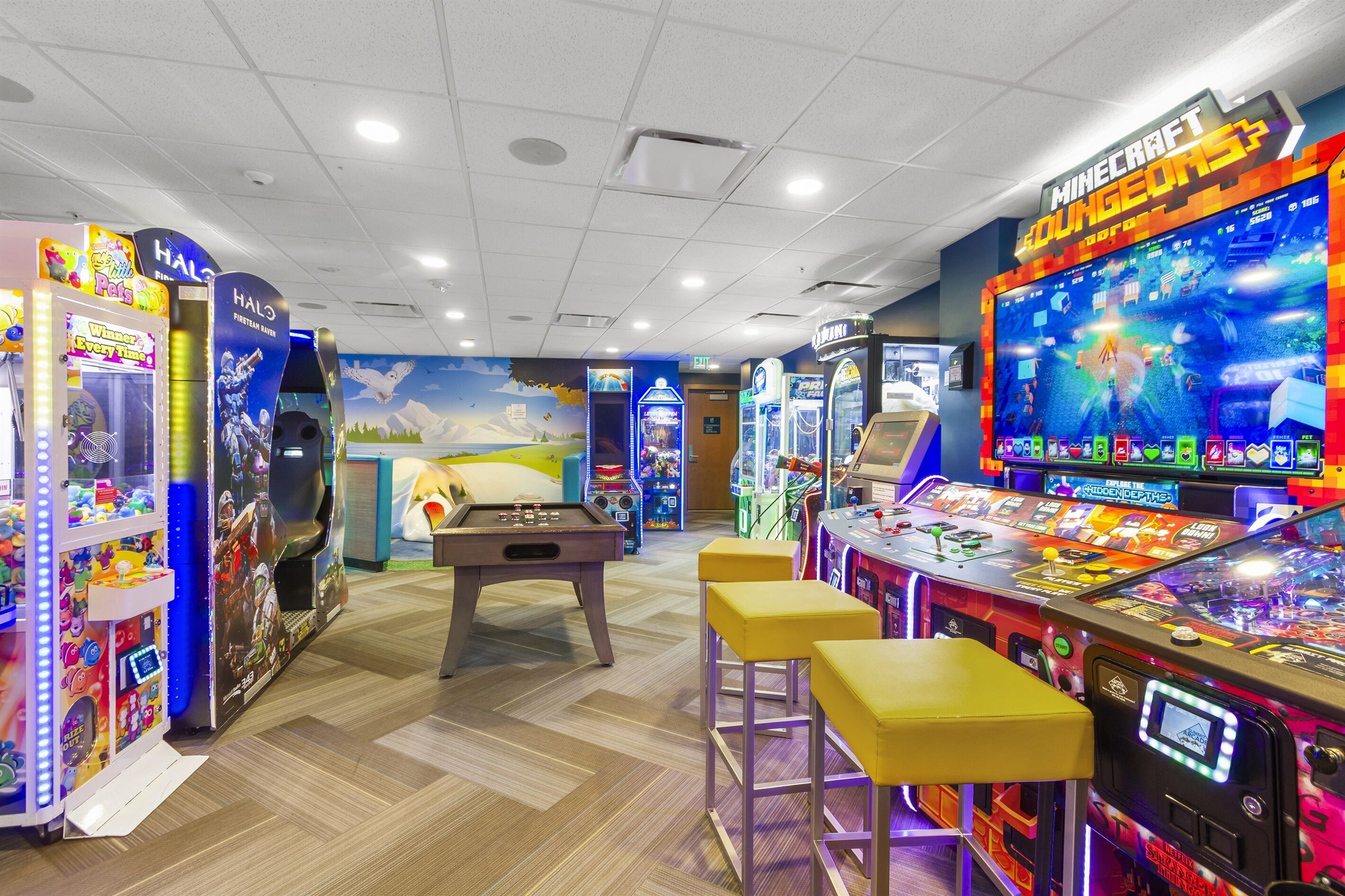 An indoor gaming area with various arcade machines, a pool table, and a large screen displaying a game.