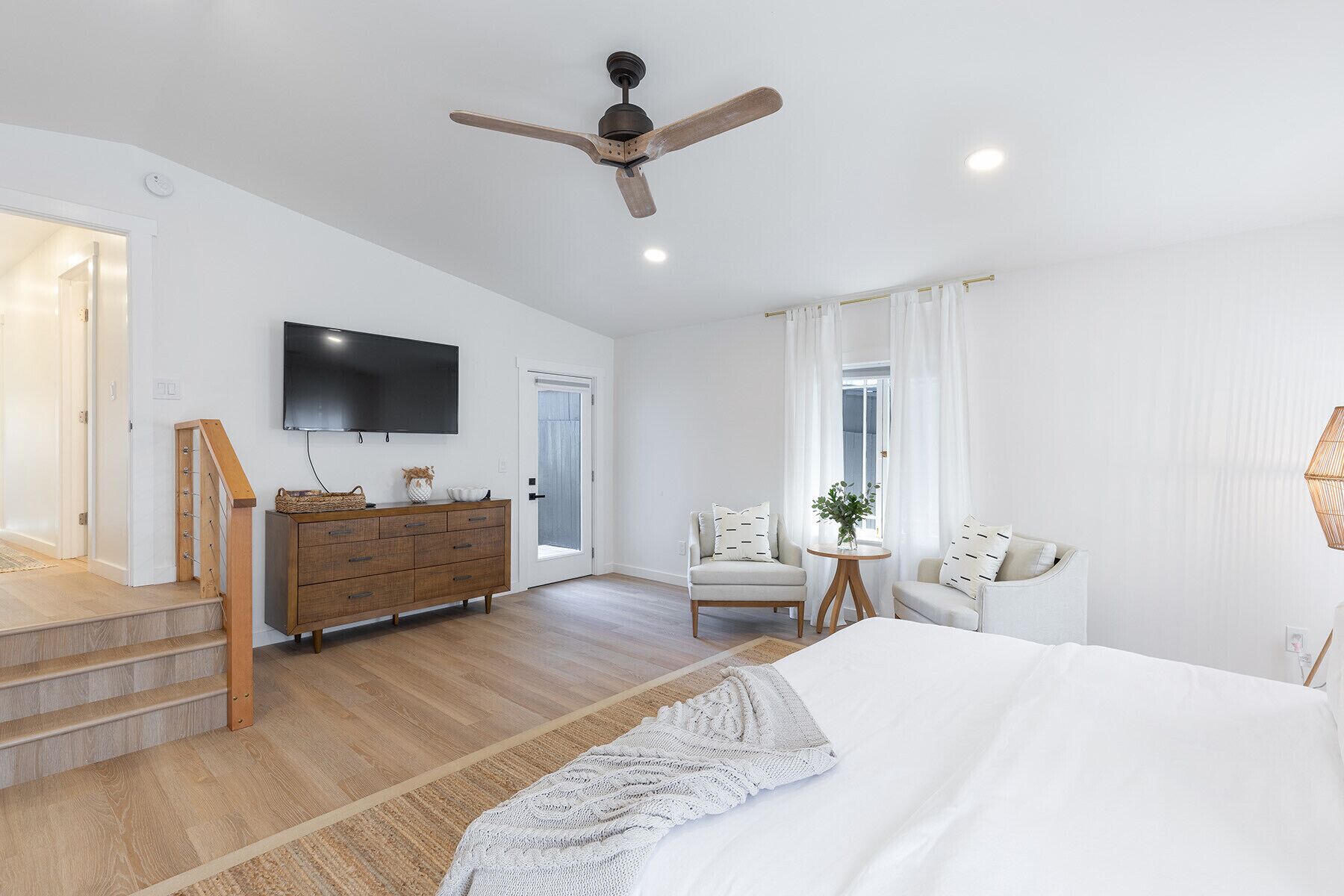 Une chambre spacieuse avec un grand lit, une commode en bois, une télévision, un ventilateur de plafond et une petite table sur laquelle est posée une plante.