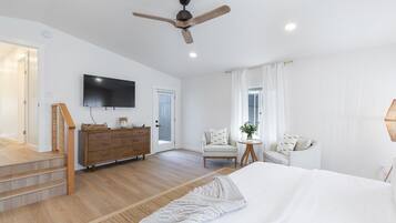 Une chambre spacieuse avec un grand lit, une commode en bois, une télévision, un ventilateur de plafond et une petite table sur laquelle est posée une plante.