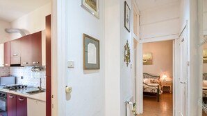 Un pasillo estrecho con una cocina a un lado y un dormitorio al otro. La cocina tiene estufa, horno y armarios. El dormitorio tiene una cama, mesita de noche y lámpara.