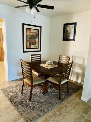 A dining area with a round table, four wooden chairs, a ceiling fan, and two framed artworks on the walls.