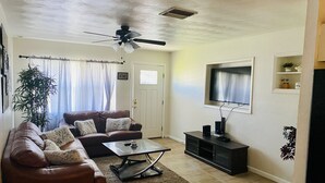 A living room with a brown leather sofa, a glass coffee table, a ceiling fan, a TV on the wall, and a door with a 'HOME' sign. - Cozy relaxation home in the heart of Cocoa (Cocoa)
