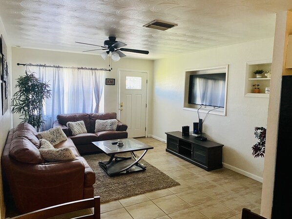 A living room with a brown leather sofa, a glass coffee table, a ceiling fan, a TV on the wall, and a door with a 'HOME' sign.