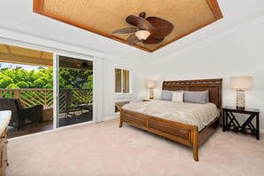 Un dormitorio con una cama grande, dos mesitas de noche con lámparas, un ventilador de techo y un ventanal corredero que da a un balcón con mobiliario exterior.