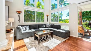 A spacious living room with a sectional sofa, a glass coffee table, and large windows offering a view of palm trees. - Hilton Pool Pass 2026 and Golf Discounts. Waikoloa Beach Villas B1 (Waikoloa Beach Resort)
