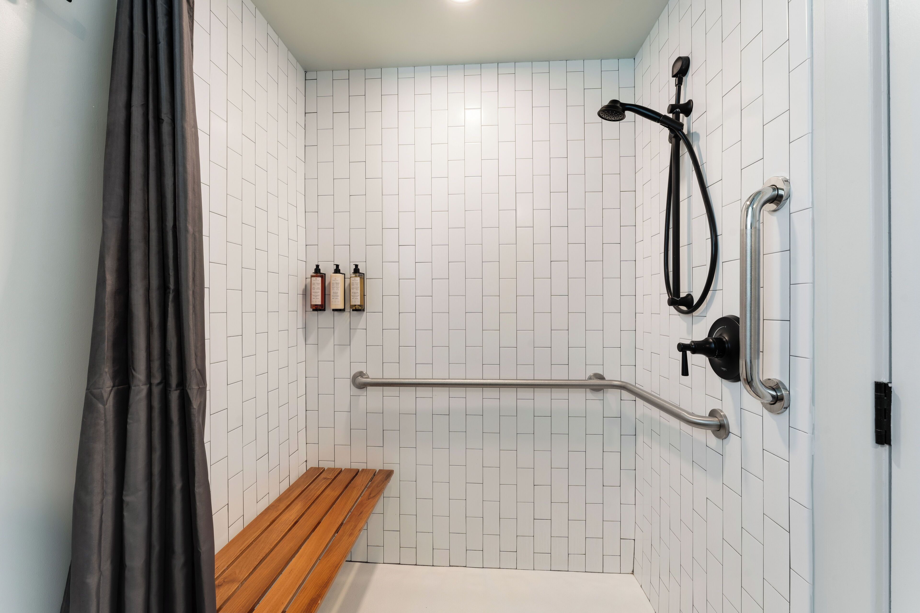 A shower area with white tiled walls, a wooden bench, a grab bar, and a showerhead.