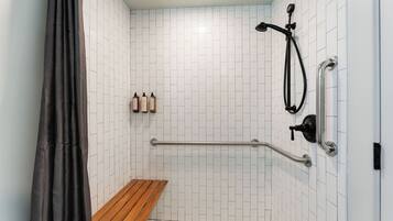 Un área de ducha con paredes de azulejos blancos, un banco de madera, una barra de apoyo y un cabezal de ducha.