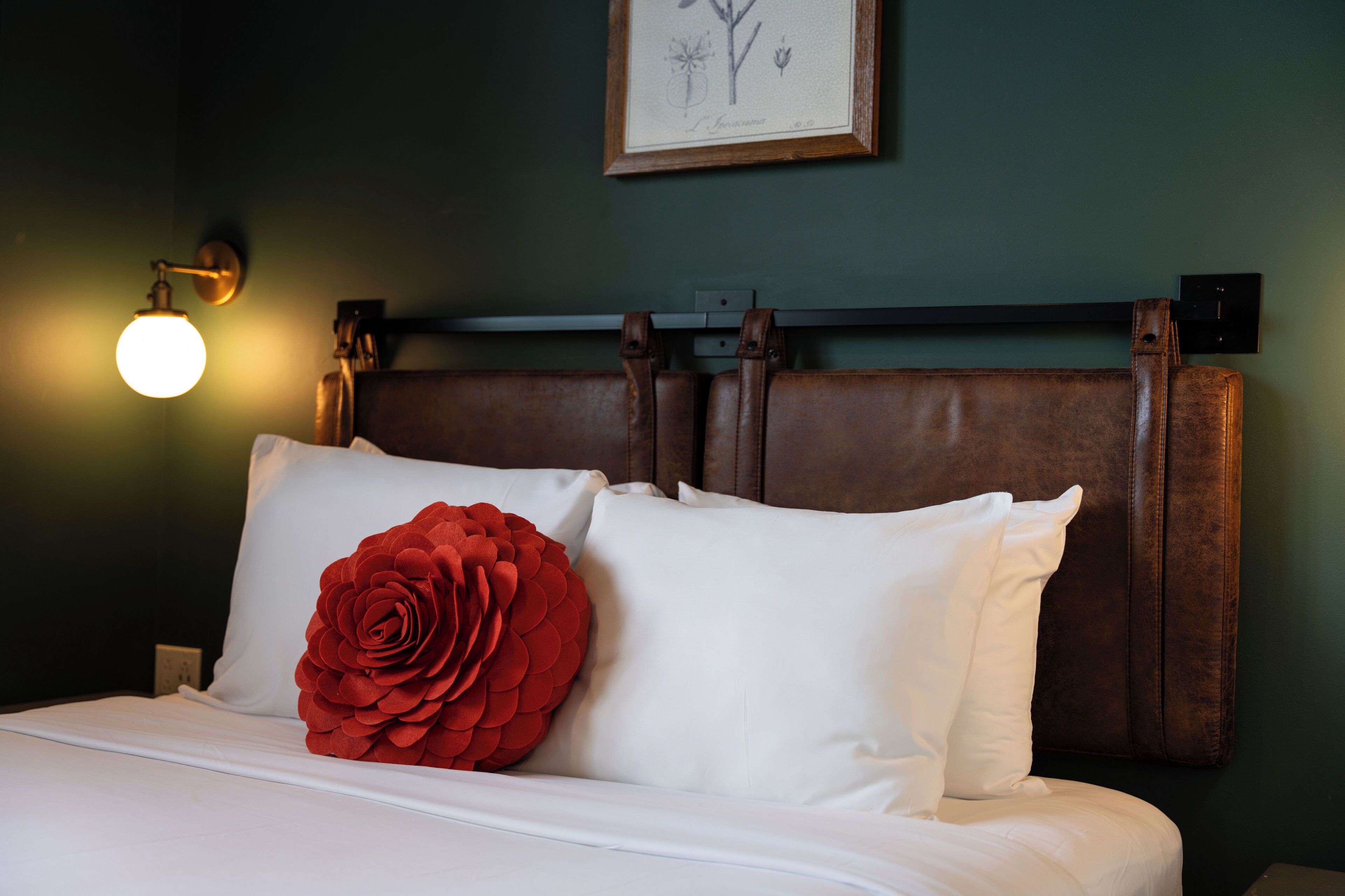 Una cama impecable con sábanas blancas y un cojín rojo con forma de rosa, una lámpara de pared y una ilustración botánica enmarcada.